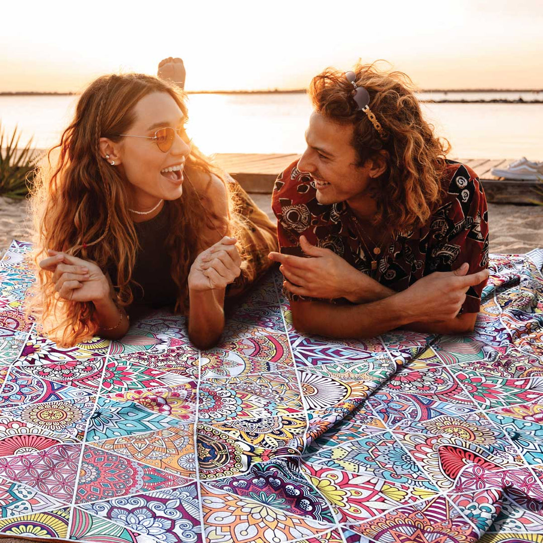 Happy hipster couple enjoy beach at sunset on oversized sand free towel.