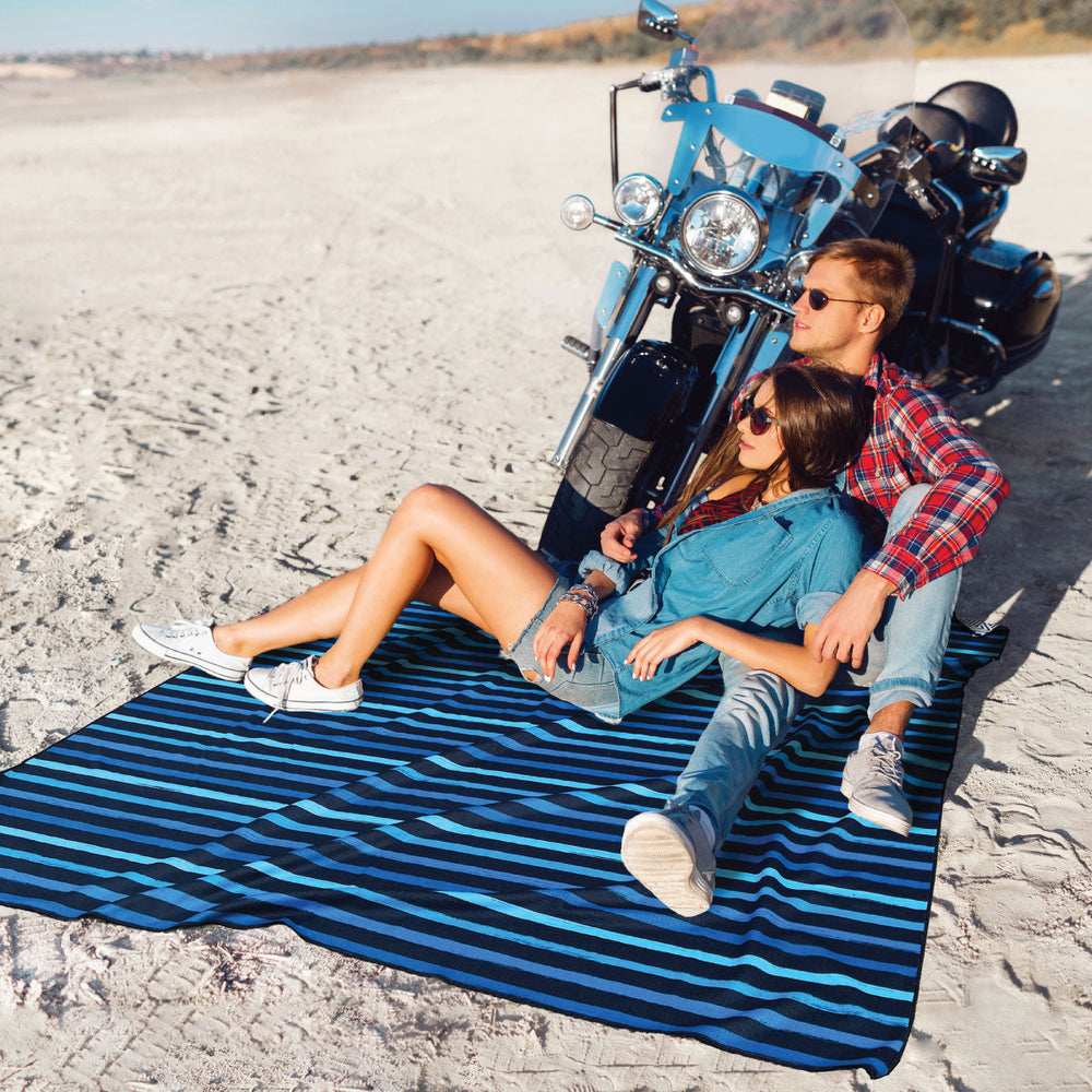 women leaning on boyfriend both are sitting on oversized beach towel at coast with motorbike in the background. 
